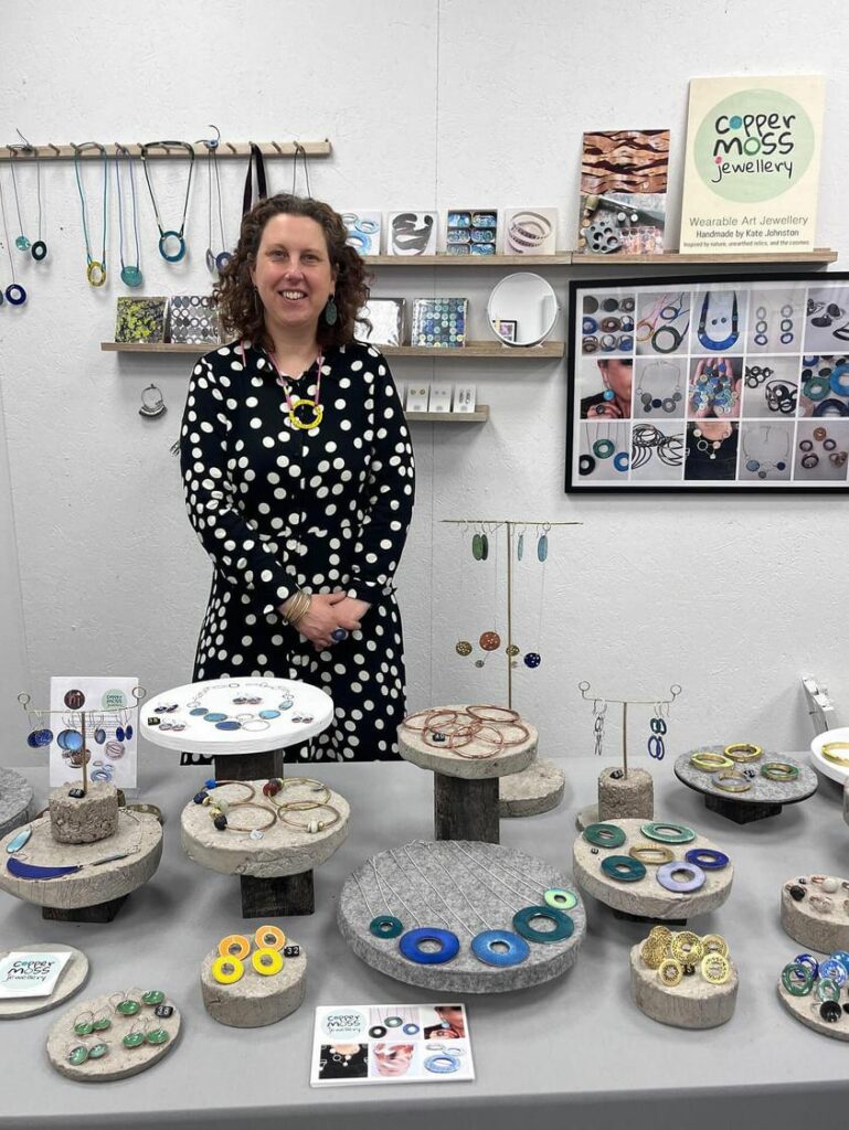 Kate - 5ft 6 female with curly brown hair, wearing a black dress with white spots. Smiling behind a display of her jewellery