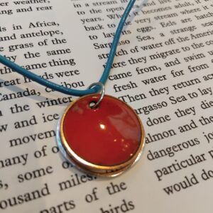 Bright orange enamelled domed circle with silver orbit hanging on marine blue leather. displayed against an open book.